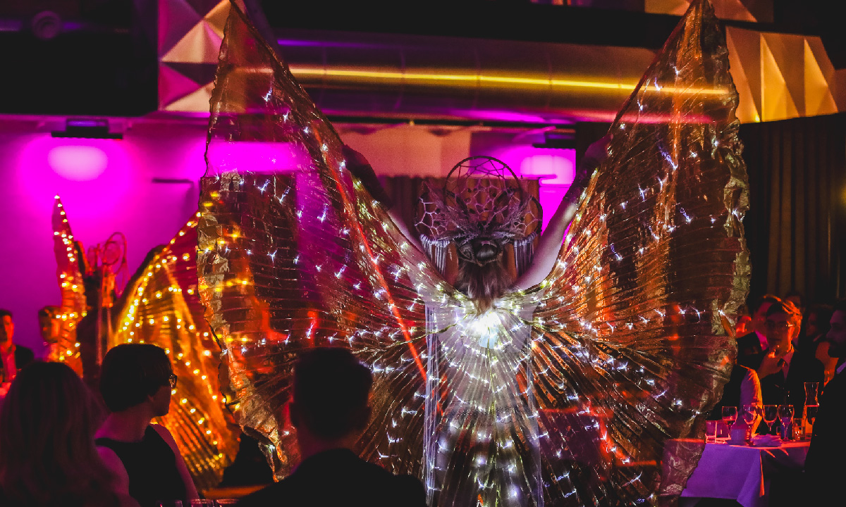 Performer with glowing wings entertaining seated guests during a corporate event at Linköping Konsert & Kongress, in a venue lit with colorful lighting.